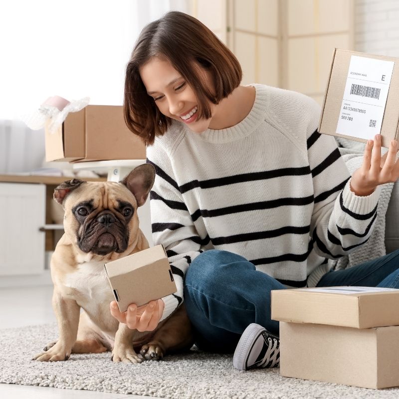 A woman and a French bulldog sit on the floor surrounded by cardboard boxes